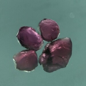 May include: Four small, dark purple gemstones, possibly amethysts, are arranged on a light blue surface.