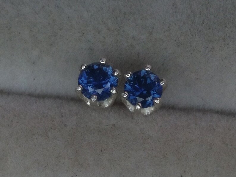 Puede incluir: Un par de pendientes de plata con piedras preciosas azules. Las piedras preciosas son redondas y tienen un corte facetado.
