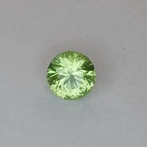 Puede incluir: Una gema redonda, facetada, de color verde claro. La gema tiene un corte brillante con múltiples facetas que reflejan la luz. El color es un verde amarillento pálido y la piedra parece transparente. La piedra preciosa está sobre un fondo neutro.
