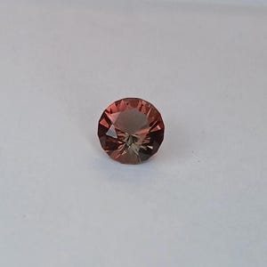May include: A faceted, round gemstone with a blend of reddish-brown and olive-green hues. The gem is set against a plain white background, showcasing its intricate cut and color variations. The gemstone's facets reflect light, creating a sparkling effect.