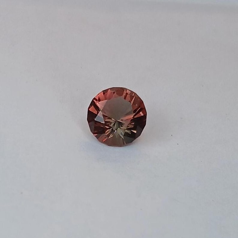 May include: A faceted, round gemstone with a blend of reddish-brown and olive-green hues. The gem is set against a plain white background, showcasing its intricate cut and color variations. The gemstone's facets reflect light, creating a sparkling effect.