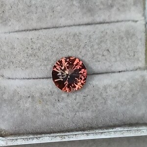 May include: A round, faceted gemstone with a reddish-orange hue. The gem is set against a gray, textured background, showcasing its intricate internal structure and brilliance. The gemstone's facets catch the light, creating a sparkling effect.