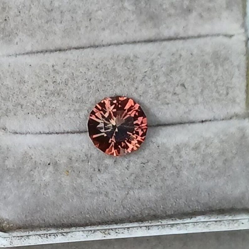 May include: A round, faceted gemstone with a reddish-orange hue. The gem is set against a gray, textured background, showcasing its intricate internal structure and brilliance. The gemstone's facets catch the light, creating a sparkling effect.