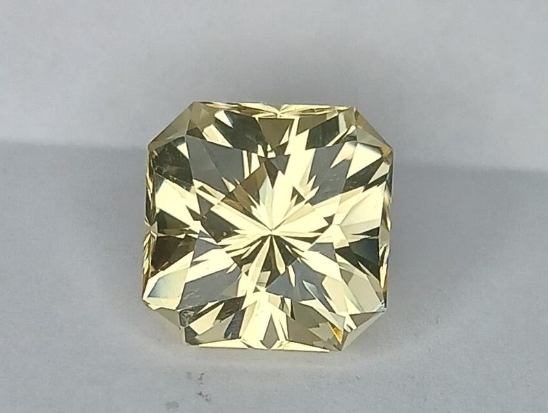 May include: A yellow gemstone with a square shape and a faceted cut. The gemstone has a star-like pattern in the centre.