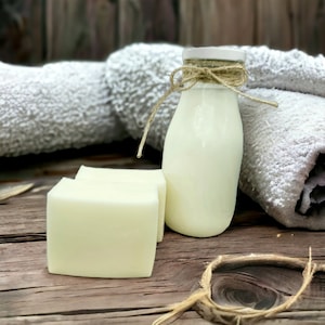 Op de afbeelding: Twee witte stukken zeep en een glazen fles melk op een houten oppervlak met een witte handdoek op de achtergrond.