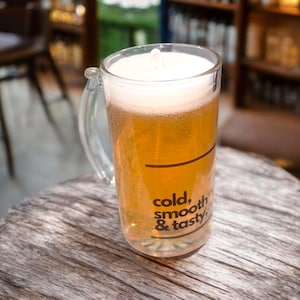 May include: A clear glass beer mug filled with a light amber liquid and topped with white foam. The mug has a handle and the words "cold, smooth & tasty" are printed on the side. The mug sits on a wooden table.