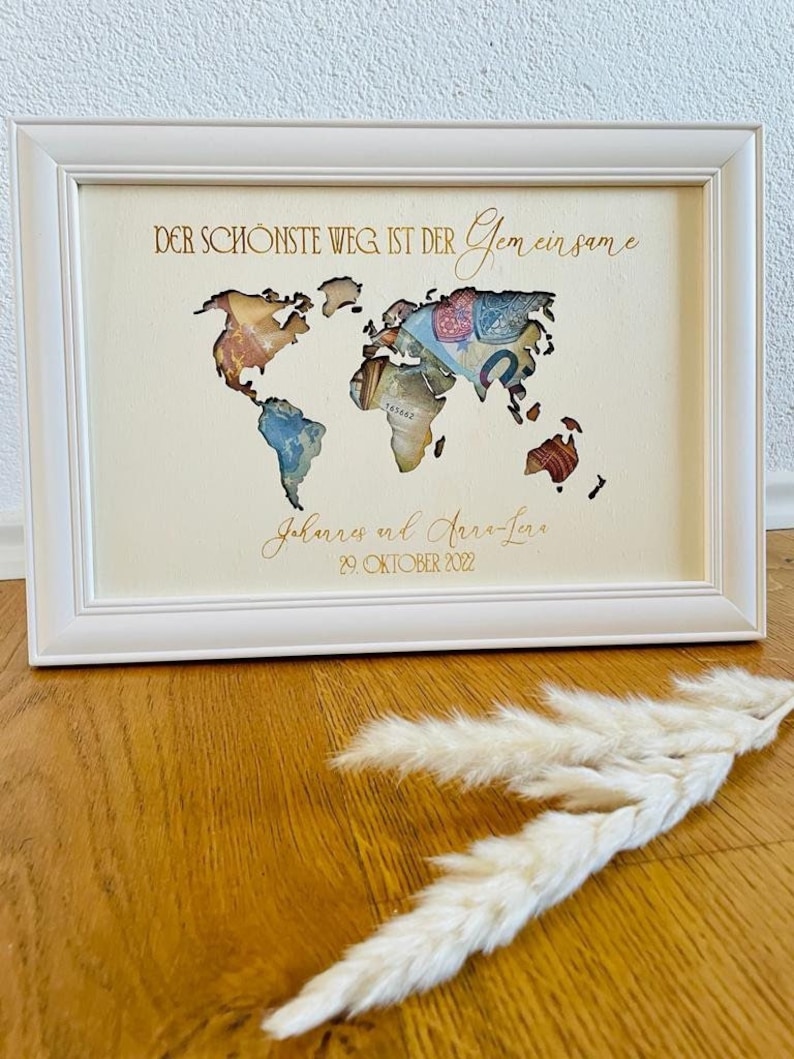 May include: A white framed picture with a world map cutout, filled with money. The text "Der sch&ouml;nste Weg ist der gemeinsame" is at the top, and "Johannes and Anna-Lena 29. Oktober 2022" is at the bottom.