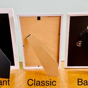 May include: Three white picture frames with different styles of easel backs. The frame on the left has a black easel back, the middle frame has a brown easel back, and the frame on the right has a black easel back. The frames are labeled "Elegant", "Classic", and "Basic" from left to right.