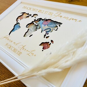 May include: A white wooden frame with a cutout world map design. The map is filled with currency bills. The text "Der sch&ouml;nste Weg ist der gemeinsame" and "Juliane und Jan-Luca 29. Oktober 2022" is written in gold lettering.