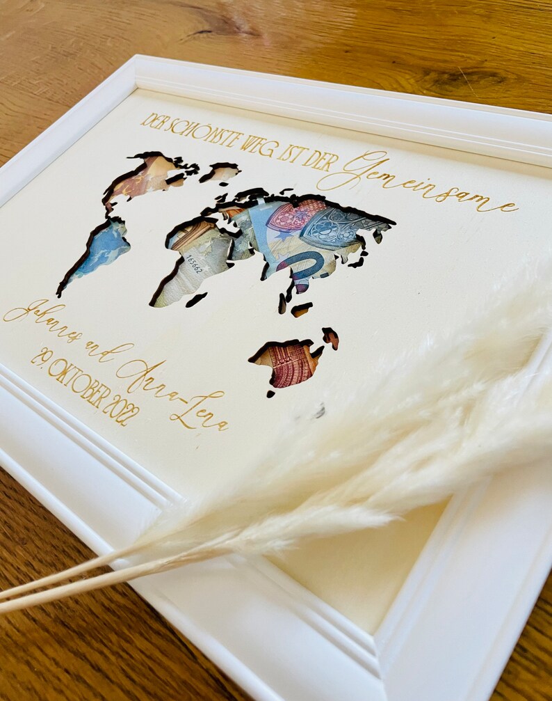 May include: A white wooden frame with a cutout world map design. The map is filled with currency bills. The text "Der sch&ouml;nste Weg ist der gemeinsame" and "Juliane und Jan-Luca 29. Oktober 2022" is written in gold lettering.
