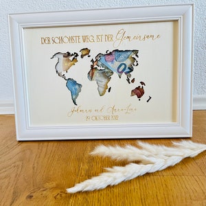 May include: A white wooden frame with a cutout world map. The map is filled with money. The text "DER SCH&Ouml;NSTE WEG IST DER Gemeinsame" is at the top of the frame. The text "Johannes und Anna-Lena 29. OKTOBER 2022" is at the bottom of the frame.