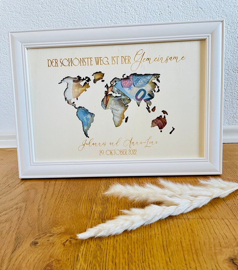 May include: A white wooden frame with a cutout world map. The map is filled with money. The text "DER SCH&Ouml;NSTE WEG IST DER Gemeinsame" is at the top of the frame. The text "Johannes und Anna-Lena 29. OKTOBER 2022" is at the bottom of the frame.