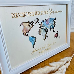 May include: A white framed world map with a gold inscription that reads "Der sch&ouml;nste Weg ist der gemeinsame" and "Johannes and Anna-Lena 29. Oktober 2022". The map is made of paper and has a cutout design.
