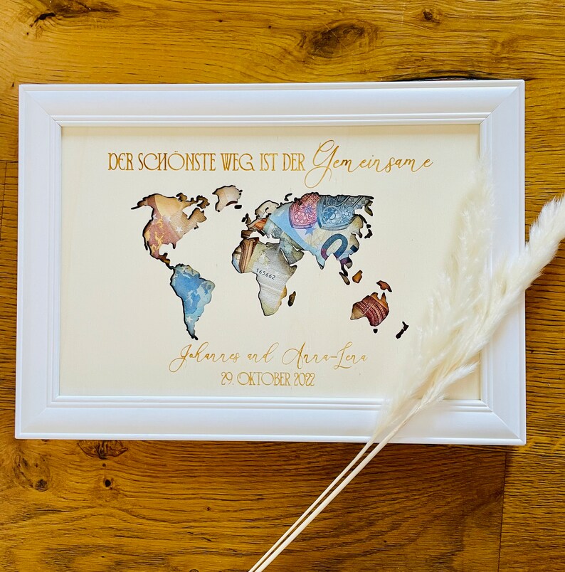 May include: A white picture frame with a gold inscription that reads "DER SCH&Ouml;NSTE WEG IST DER Gemeinsame Johannes und Anna-Lena 29. OKTOBER 2022". The frame contains a world map cutout with a background of currency bills.