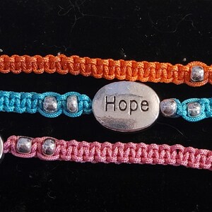 May include: Three friendship bracelets with silver oval charms that say "Love", "Hope", and "Peace". The bracelets are made with orange, blue, and pink cord.