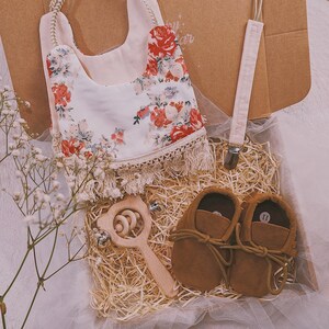 K&ouml;nnte beinhalten: Ein Baby-Geschenkset in einer Box mit einem floralen L&auml;tzchen, einem Schnullerclip, braunen Baby-Lederschuhen und einer Holzrassel. Das L&auml;tzchen hat einen cremefarbenen Hintergrund mit roten und rosa Blumenmustern. Die Schuhe sind Gr&ouml;&szlig;e 11.