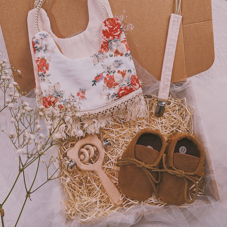 K&ouml;nnte beinhalten: Ein Baby-Geschenkset in einer Box mit einem floralen L&auml;tzchen, einem Schnullerclip, braunen Baby-Lederschuhen und einer Holzrassel. Das L&auml;tzchen hat einen cremefarbenen Hintergrund mit roten und rosa Blumenmustern. Die Schuhe sind Gr&ouml;&szlig;e 11.