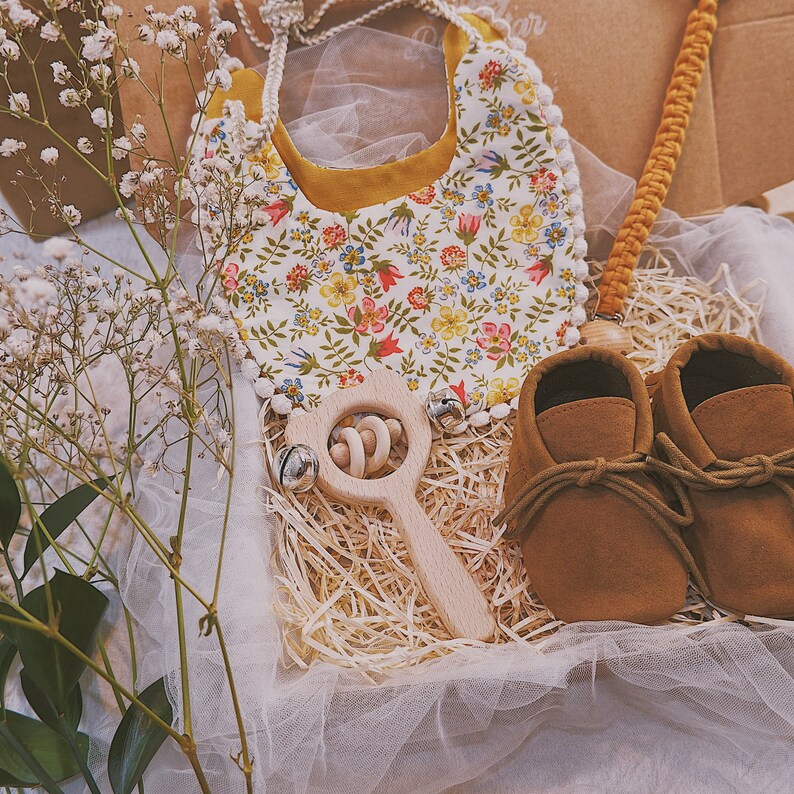 K&ouml;nnte beinhalten: Ein Baby-Geschenkset mit einem L&auml;tzchen mit floralem Muster und gelbem Rand, braunen Babyschuhen aus Wildleder, einer Holzrassel und einem Schnullerclip. Die Artikel sind in einer Geschenkbox mit Stroh und T&uuml;ll angeordnet.