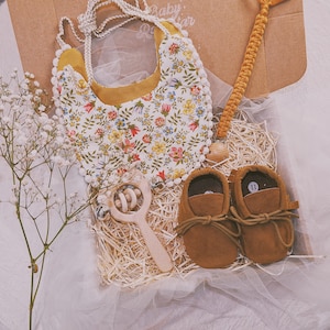 K&ouml;nnte beinhalten: Ein Baby-Geschenkset in einer Box mit einem floralen L&auml;tzchen, einer Holzrassel, einem Schnullerclip und braunen Wildleder-Babyschuhen. Das L&auml;tzchen hat einen wei&szlig;en Hintergrund mit bunten Blumen und gelbem Besatz. Die Schuhe sind Gr&ouml;&szlig;e 11.