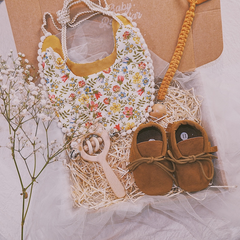 K&ouml;nnte beinhalten: Ein Baby-Geschenkset in einer Box mit einem floralen L&auml;tzchen, einer Holzrassel, einem Schnullerclip und braunen Wildleder-Babyschuhen. Das L&auml;tzchen hat einen wei&szlig;en Hintergrund mit bunten Blumen und gelbem Besatz. Die Schuhe sind Gr&ouml;&szlig;e 11.
