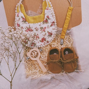 K&ouml;nnte beinhalten: Ein Baby-Geschenkset in einer Pappschachtel, einschlie&szlig;lich einer floralen L&auml;tzchen, einer Holzrassel, einem Schnullerclip und braunen Wildleder-Babyschuhen. Das L&auml;tzchen hat einen wei&szlig;en Hintergrund mit roten und rosa Rosen. Die Schuhe sind Gr&ouml;&szlig;e 11.