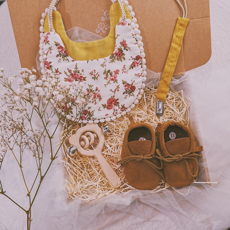K&ouml;nnte beinhalten: Ein Baby-Geschenkset in einer Pappschachtel, einschlie&szlig;lich einer floralen L&auml;tzchen, einer Holzrassel, einem Schnullerclip und braunen Wildleder-Babyschuhen. Das L&auml;tzchen hat einen wei&szlig;en Hintergrund mit roten und rosa Rosen. Die Schuhe sind Gr&ouml;&szlig;e 11.
