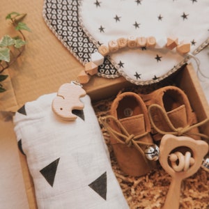 Peut inclure: Une bo&icirc;te-cadeau remplie d'articles pour b&eacute;b&eacute;, dont une couverture en mousseline &agrave; motifs blancs et noirs, une paire de chaussures de b&eacute;b&eacute; en cuir marron, un hochet en bois et une t&eacute;tine en bois en forme d'&eacute;l&eacute;phant attach&eacute;e &agrave; un collier de perles avec le nom "Darcie" orthographi&eacute;.