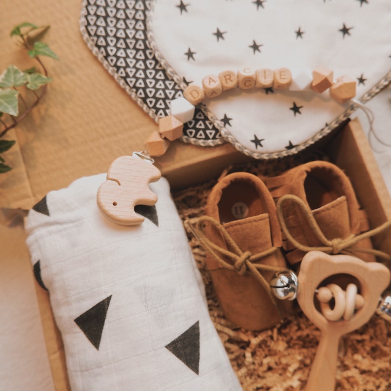 Peut inclure: Une bo&icirc;te-cadeau remplie d'articles pour b&eacute;b&eacute;, dont une couverture en mousseline &agrave; motifs blancs et noirs, une paire de chaussures de b&eacute;b&eacute; en cuir marron, un hochet en bois et une t&eacute;tine en bois en forme d'&eacute;l&eacute;phant attach&eacute;e &agrave; un collier de perles avec le nom "Darcie" orthographi&eacute;.