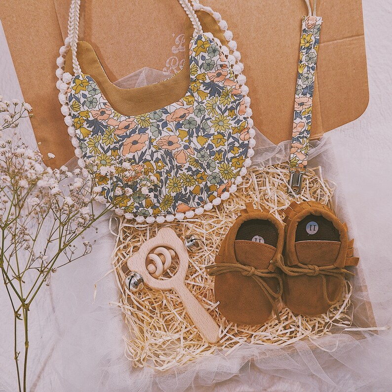 K&ouml;nnte beinhalten: Ein Baby-Geschenkset in einer Box, einschlie&szlig;lich einer floralen L&auml;tzchen mit wei&szlig;em Pomponbesatz, einem passenden Schnullerclip, braunen Wildleder-Babyschuhen und einer Holzrassel. L&auml;tzchen und Clip haben ein farbenfrohes Blumenmuster.