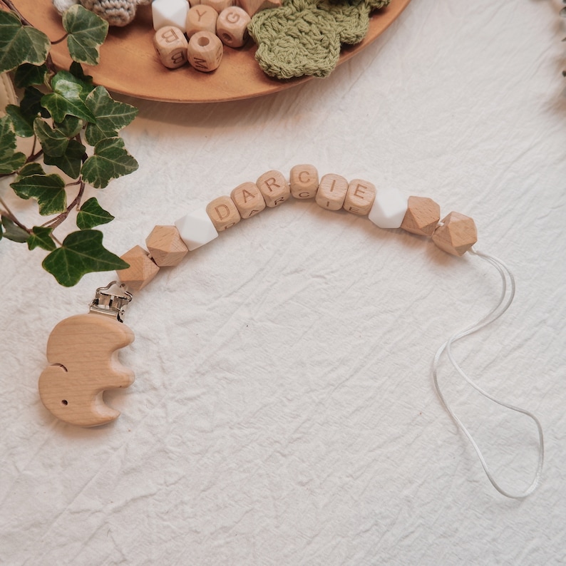 Peut inclure: Un hochet en bois pour b&eacute;b&eacute; avec le nom "DARCIE" &eacute;crit en lettres blanches. Le hochet est muni d'un &eacute;l&eacute;phant en bois attach&eacute; &agrave; un clip.
