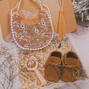 K&ouml;nnte beinhalten: Ein Baby-Geschenkset in einem Karton. Es enth&auml;lt eine L&auml;tzchen mit Blumenmuster und wei&szlig;er Bord&uuml;re, einen Schnullerclip, braune Babyschuhe aus Wildleder und eine Holzrassel mit Gl&ouml;ckchen. Das L&auml;tzchen hat ein orangefarbenes Muster mit wei&szlig;en Blumen.