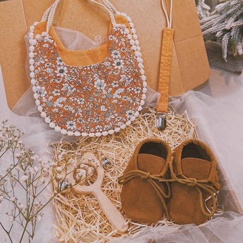 K&ouml;nnte beinhalten: Ein Baby-Geschenkset in einem Karton. Es enth&auml;lt eine L&auml;tzchen mit Blumenmuster und wei&szlig;er Bord&uuml;re, einen Schnullerclip, braune Babyschuhe aus Wildleder und eine Holzrassel mit Gl&ouml;ckchen. Das L&auml;tzchen hat ein orangefarbenes Muster mit wei&szlig;en Blumen.
