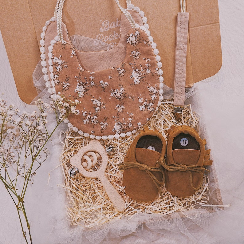 K&ouml;nnte beinhalten: Baby-Geschenkset in einer Box mit floralem L&auml;tzchen, braunen Wildleder-Babyschuhen, einer Holzrassel und einem Schnullerclip. Das L&auml;tzchen hat ein Blumenmuster mit wei&szlig;em Besatz. Die Babyschuhe sind Gr&ouml;&szlig;e II. Die Box ist mit Stroh gef&uuml;llt.