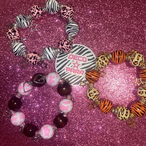 Könnte beinhalten: Drei Armbänder aus bunten Holzperlen mit verschiedenen Mustern. Die Armbänder haben einen Zebra-, einen Kuh- und einen Leopardenprint. Die Armbänder haben silberne Charms in Form von Sternen und Herzen.