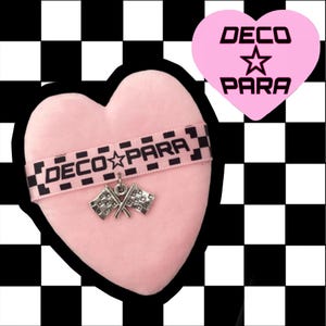Op de afbeelding: Een roze hartvormig object met een zwart-wit geruit lint met de tekst "DECO PARA". Een zilveren bedel van twee geblokte vlaggen hangt aan het lint. Een roze hartvormige afbeelding met de woorden "DECO PARA" staat in de rechterbovenhoek.