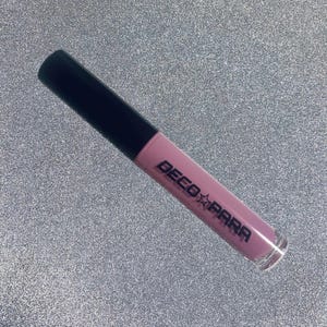 Può includere: Un tubetto di lucidalabbra malva con tappo nero. Le parole "DECO PARA" sono stampate in nero sul tubetto. Il lucidalabbra è su uno sfondo glitterato argentato.