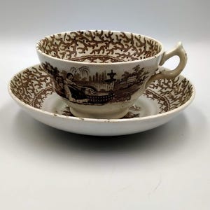 Puede incluir: Un juego de taza y platillo vintage con un patrón floral marrón y blanco. La taza tiene un asa y el platillo tiene un borde festoneado.