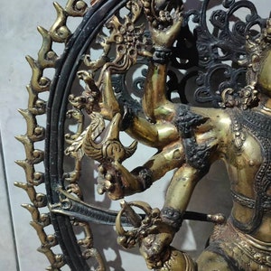 Op de afbeelding: Een gouden metalen sculptuur van een godheid met meerdere armen, staand in een cirkelvormig frame met ingewikkeld zwart metaalwerk. De godheid is versierd met sieraden en heeft een woeste uitdrukking.