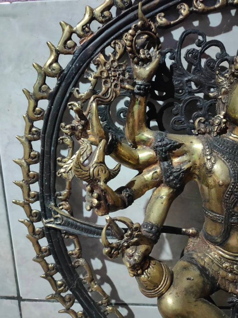 Op de afbeelding: Een gouden metalen sculptuur van een godheid met meerdere armen, staand in een cirkelvormig frame met ingewikkeld zwart metaalwerk. De godheid is versierd met sieraden en heeft een woeste uitdrukking.
