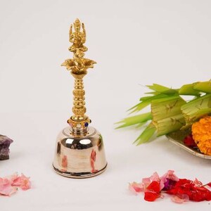 Op de afbeelding: Een zilveren bel met een gouden top met een godheidfiguur. De bel is omringd door roze en rode bloemblaadjes, paarse kristallen en een bord met groene bladeren, gele bloemen en gouden munten.