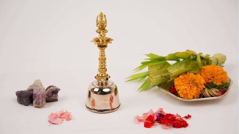 Op de afbeelding: Een zilveren bel met een gouden top met een godheidfiguur. De bel is omringd door roze en rode bloemblaadjes, paarse kristallen en een bord met groene bladeren, gele bloemen en gouden munten.