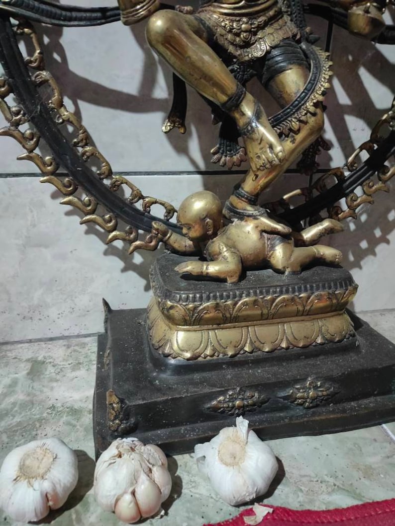 Op de afbeelding: Een gouden beeld van een dansende figuur met een kind eronder. Het beeld staat op een zwarte basis met gouden accenten. Het beeld is omringd door drie witte knoflookbollen.
