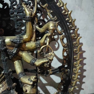 Op de afbeelding: Een gouden metalen sculptuur van een godheid met meerdere armen, die verschillende voorwerpen vasthoudt. Het beeld is omgeven door een zwart metalen frame met ingewikkelde ontwerpen.
