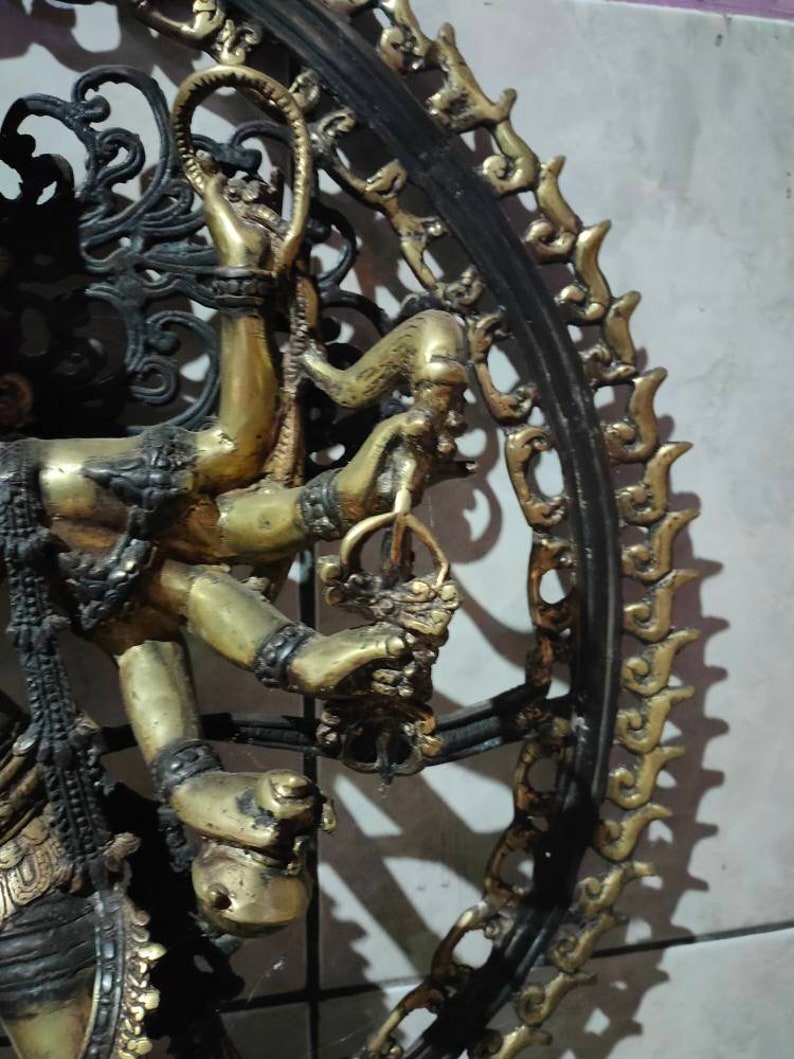 Op de afbeelding: Een gouden metalen sculptuur van een godheid met meerdere armen, die verschillende voorwerpen vasthoudt. Het beeld is omgeven door een zwart metalen frame met ingewikkelde ontwerpen.