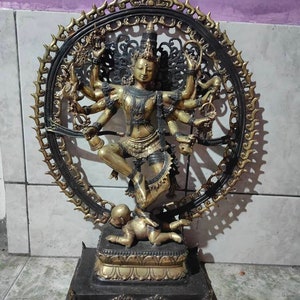 Op de afbeelding: Een gouden en zwart metalen beeld van een hindoe&iuml;stische godheid met meerdere armen, dansend op een demon. Het beeld is omgeven door een cirkelvormige halo met ingewikkelde ontwerpen. De godheid draagt een kroon en sieraden.