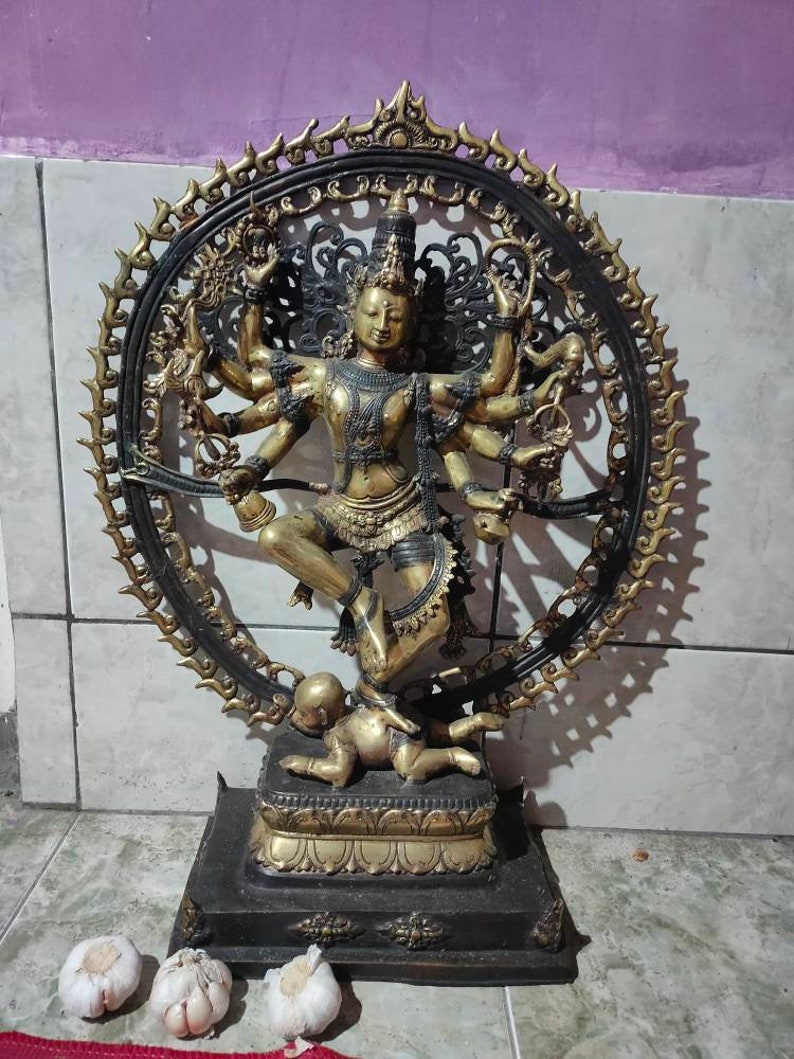 Op de afbeelding: Een gouden en zwart metalen beeld van een hindoe&iuml;stische godheid met meerdere armen, dansend op een demon. Het beeld is omgeven door een cirkelvormige halo met ingewikkelde ontwerpen. De godheid draagt een kroon en sieraden.