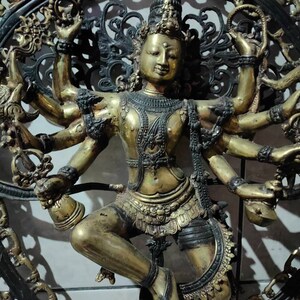 Op de afbeelding: Een gouden metalen sculptuur van een godheid met meerdere armen, die een kroon en uitgebreide sieraden draagt, dansend in een cirkelvormig frame met ingewikkeld zwart metaalwerk.