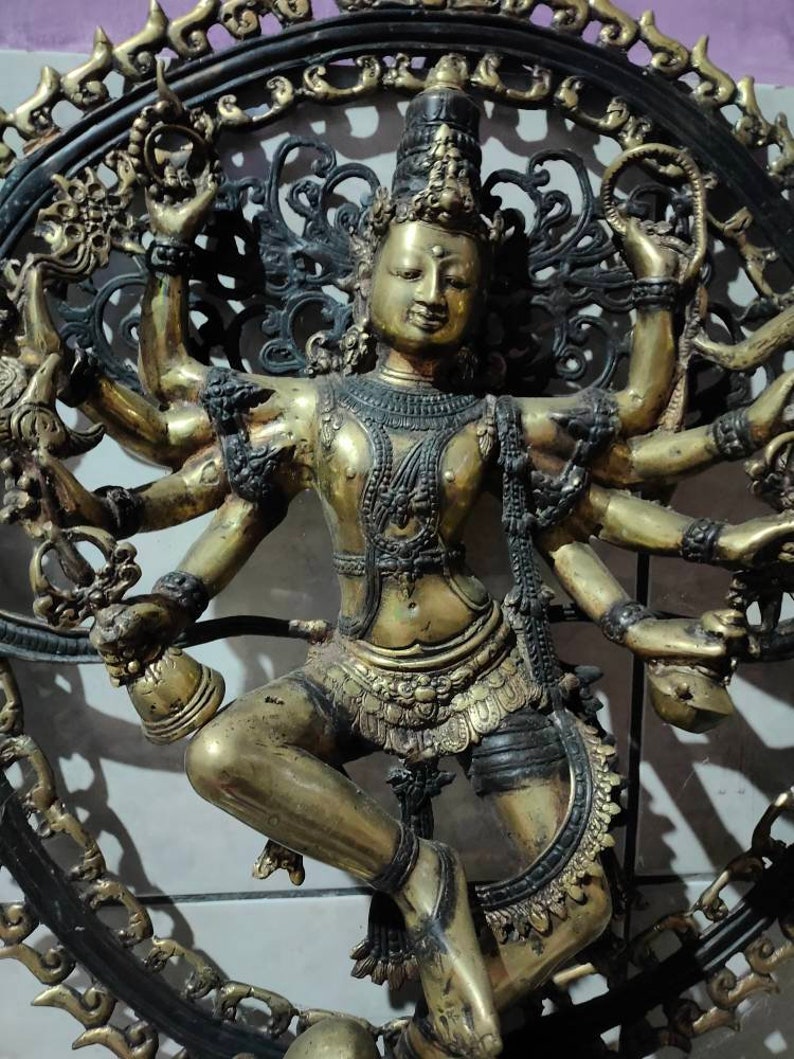 Op de afbeelding: Een gouden metalen sculptuur van een godheid met meerdere armen, die een kroon en uitgebreide sieraden draagt, dansend in een cirkelvormig frame met ingewikkeld zwart metaalwerk.