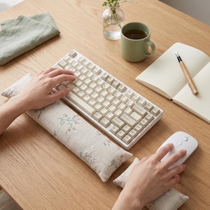 May include: A beige mechanical keyboard with matching keycaps sits on a light wood desk, next to a white mouse. Floral-patterned wrist rests and a notebook with a pen are also on the desk. A small vase of flowers and a green mug are also present.