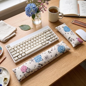 May include: A light-colored wooden desk with a white keyboard, two floral-patterned wrist rests, a cup of tea, and a small vase of flowers. A notebook with drawings and colored pencils are also on the desk.